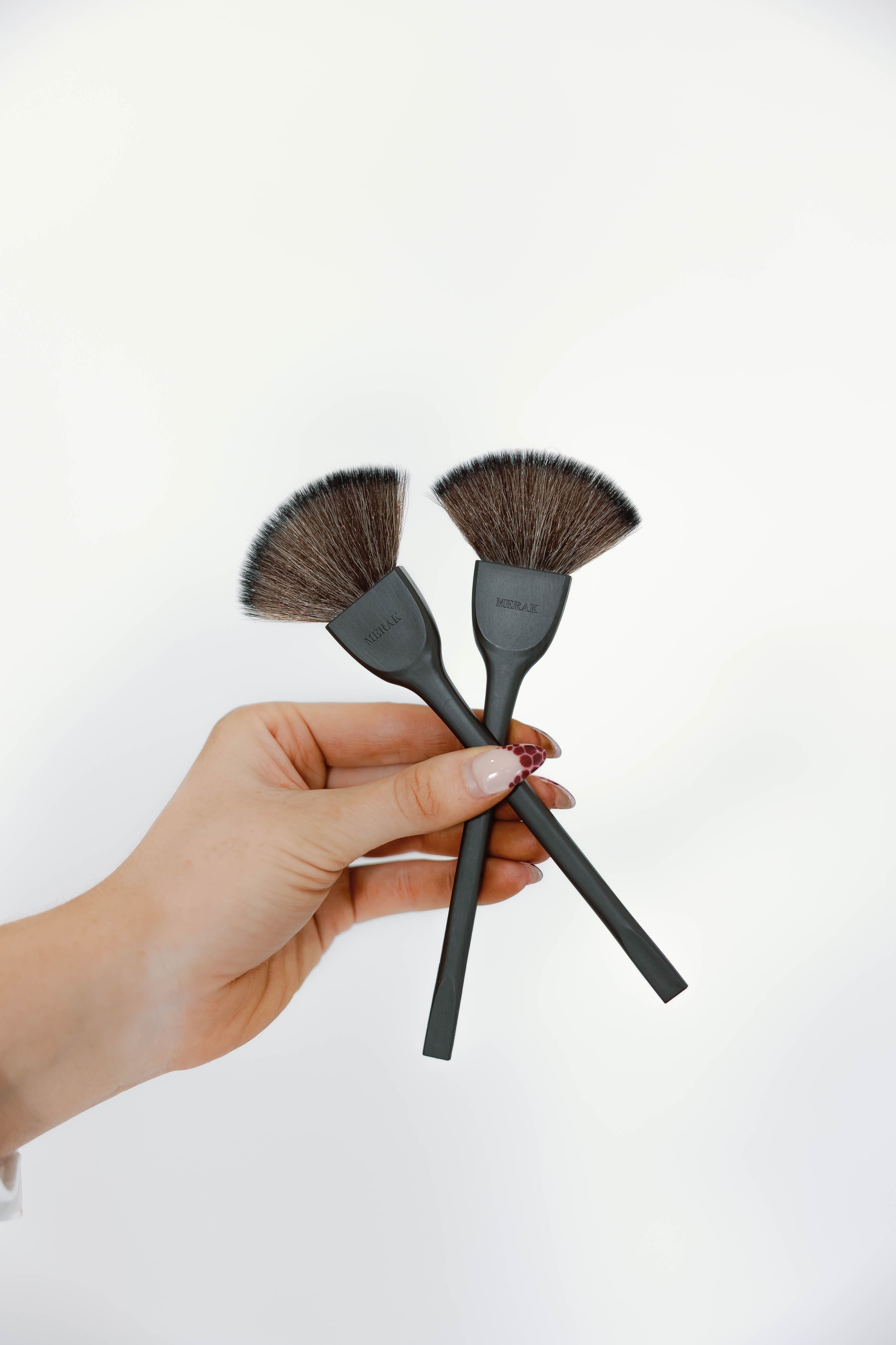 A hand holding two black makeup brushes with dark brown bristles against a plain white background.