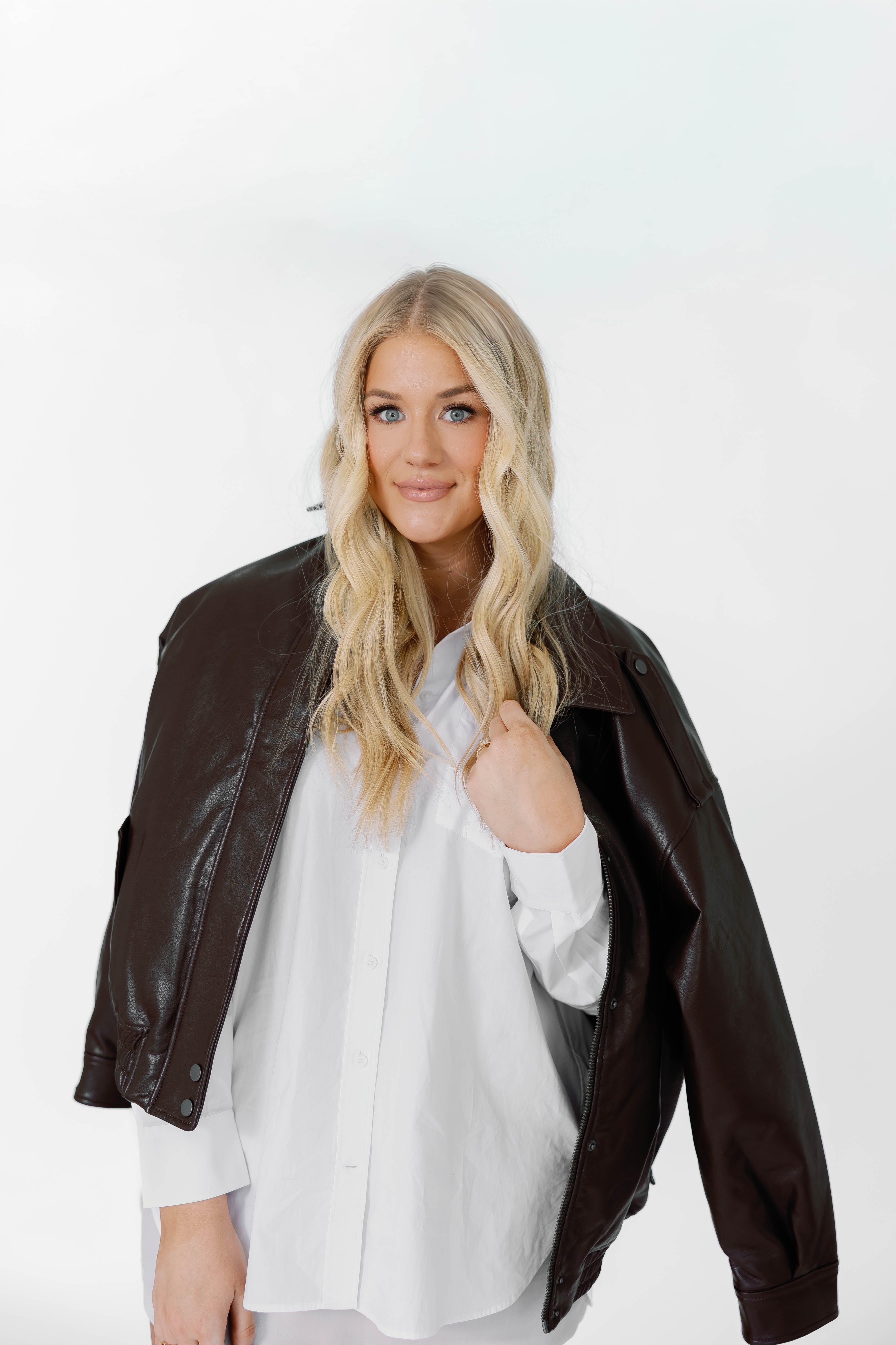 A portrait of Caroline Ply wearing a dark brown faux-leather jacket over a white button-up shirt, photographed against a clean white studio background.