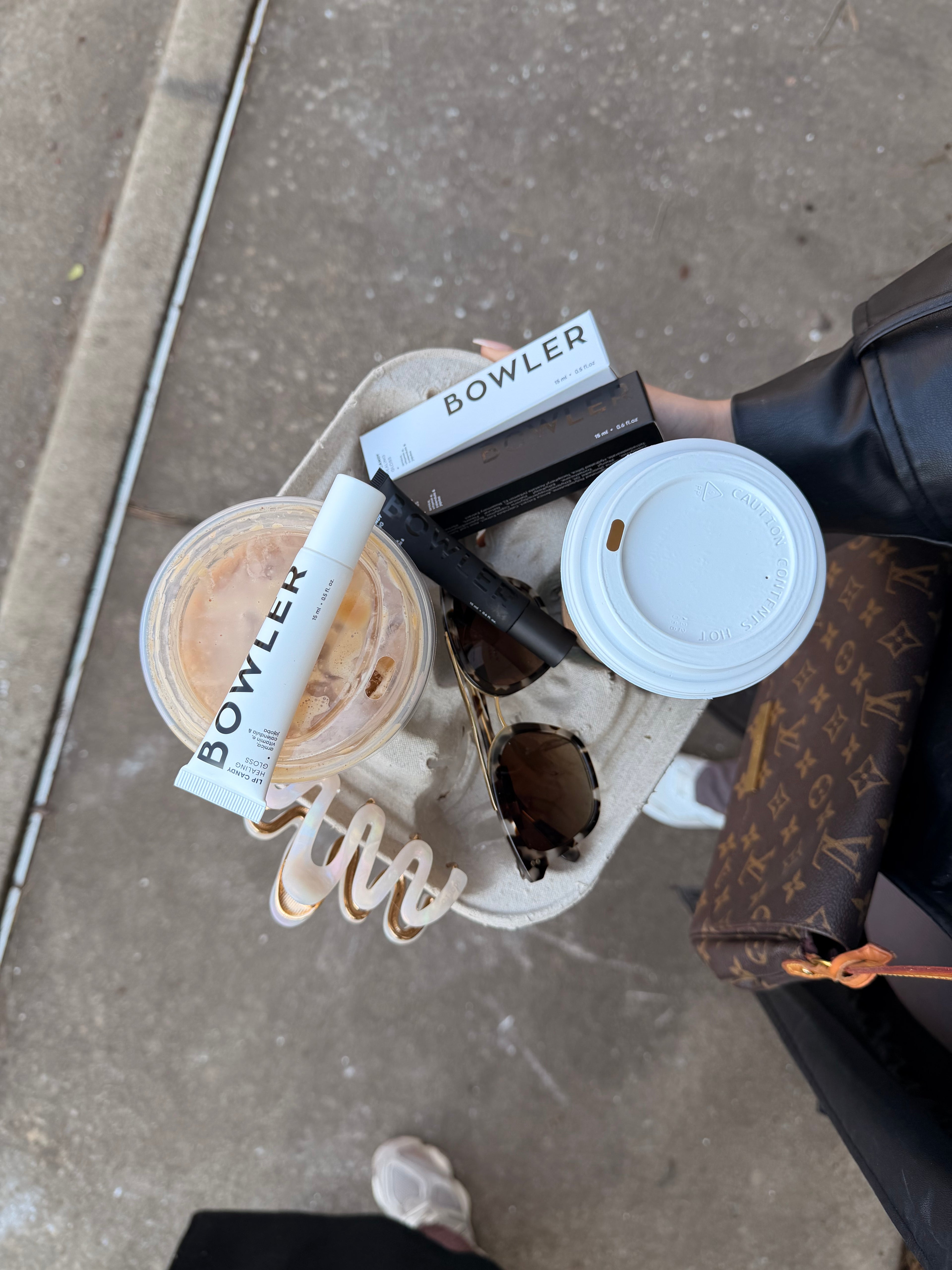 A top-down view of a person holding a beige tray with a Bowler-branded cosmetic tube, a boxed item, a black pen-like object, a white coffee lid, and a clear cup on a concrete sidewalk. A Louis Vuitton monogram bag is also visible at the right edge.