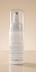 A white pump bottle with a clear cap and a white label featuring gold lettering that reads 'XTAN' and Sunless Tanning Mousse set against a warm beige background.