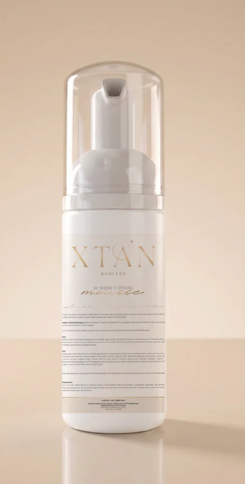 A white pump bottle with a clear cap and a white label featuring gold lettering that reads 'XTAN' and Sunless Tanning Mousse set against a warm beige background.