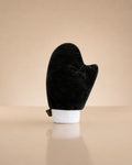 A black velour mitt with a white cuff, displayed upright on a beige reflective surface against a matching beige background.