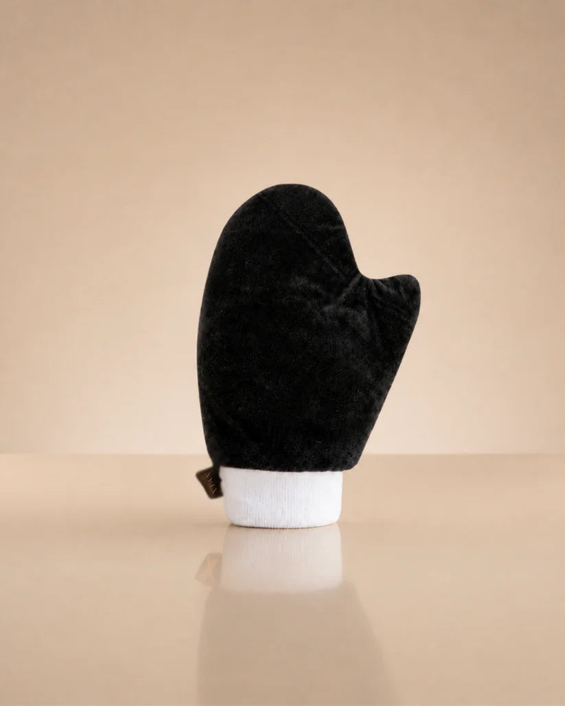 A black velour mitt with a white cuff, displayed upright on a beige reflective surface against a matching beige background.